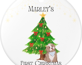 May include: A round white ceramic Christmas ornament with a gold hanging loop. It features a green Christmas tree with red bows, a star, and a dog. Text reads "Marley's First Christmas 2025".