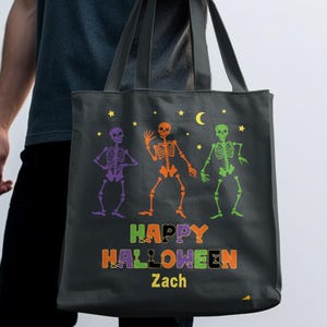 Puede incluir: Un bolso tote de Halloween gris oscuro con tres figuras de esqueleto coloridas. La bolsa tiene las palabras "HAPPY HALLOWEEN" en naranja, verde y morado, con el nombre "Zach" debajo. También son visibles estrellas amarillas y una luna creciente.