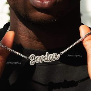 May include: A silver necklace with the name "Jordan" spelled out in a cursive font, encrusted with small, sparkling stones. The necklace is held against a dark background, with the brand name "Junaidopal" visible.
