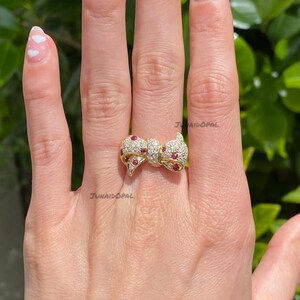 May include: A gold ring featuring a bow-shaped centerpiece adorned with small clear stones and scattered round red stones. The text JUNAIDOPAL is visible on the ring. The bow design is symmetrical with a central knot and two looped sides.