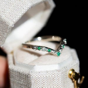 May include: A silver ring with a V-shaped design, adorned with small, bright green gemstones. The ring is presented in a white, textured jewellery box with a gold clasp, set against a black backdrop. The ring's band features a delicate, textured pattern.