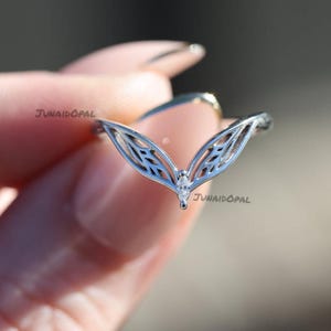 May include: A silver ring with a delicate, openwork design. The ring features a V-shaped band with a central marquise-cut gemstone. The design incorporates leaf-like patterns, creating an elegant and nature-inspired aesthetic. The ring is held up by a person.