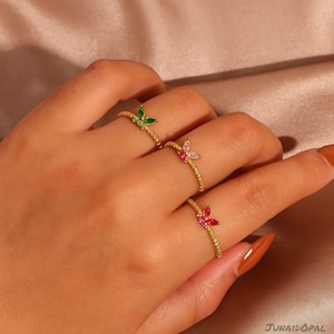 Könnte beinhalten: Drei goldfarbene Ringe, jeweils mit einem Schmetterlingsdesign. Die Schmetterlinge sind mit grünen, rosa und roten Edelsteinen verziert. Die Ringe werden auf einer Hand präsentiert, um das Design und die Farbvariationen des Schmucks hervorzuheben.