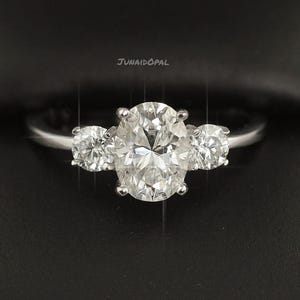 May include: A silver engagement ring featuring an oval-cut central diamond flanked by two smaller round diamonds. The ring's design is showcased against a dark backdrop, emphasizing the brilliance of the stones.