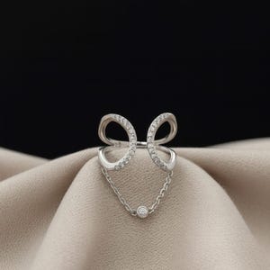 May include: A silver ring with a unique design. The ring features two curved, open-ended shapes adorned with small, clear stones. A delicate chain connects the shapes, culminating in a single, round stone. The ring is displayed on a beige fabric.