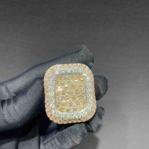 May include: A rectangular ring with a gold-colored center, surrounded by a row of small, clear stones. The outer edge of the ring is a rose gold color. The ring is held by a gloved hand.