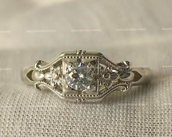May include: A silver-toned metal ring featuring a central round brilliant-cut clear gemstone set within a square frame. The band displays intricate floral engravings and openwork detailing on both sides of the central stone setting.