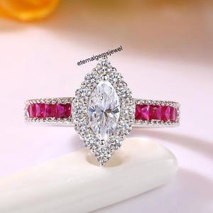Puede incluir: Un anillo de diamantes con una piedra central en forma de marquesa, rodeada por un halo de diamantes más pequeños. La banda presenta una fila de piedras preciosas rojas de corte cuadrado. El anillo está sobre una superficie blanca, con el texto "eternalgemsjewel".