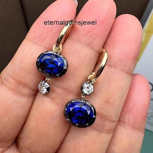 May include: Gold hoop earrings featuring oval, deep blue gemstones and small, round, clear gemstones. The earrings have a dark metal border around the blue stones. The text "eternalgemsjewel" is visible.