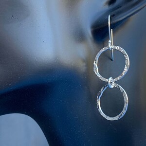 May include: A pair of silver-toned drop earrings featuring two interconnected circular rings. Each ring has a hammered, textured surface that reflects light. The earrings are suspended from a simple silver-toned hook wire for attachment.