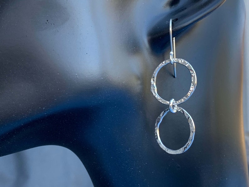 May include: A pair of silver-toned drop earrings featuring two interconnected circular rings. Each ring has a hammered, textured surface that reflects light. The earrings are suspended from a simple silver-toned hook wire for attachment.