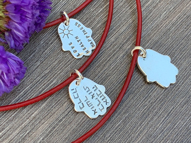 May include: Two red string bracelets with silver Hamsa hand charms. One charm has the words "Love, Health, Happiness" engraved on it, and the other charm has Hebrew writing.