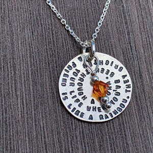 May include: Silver necklace with a round pendant that reads "The rainbow you live through is like a friend and your life is enough". The pendant has a small amber crystal charm.