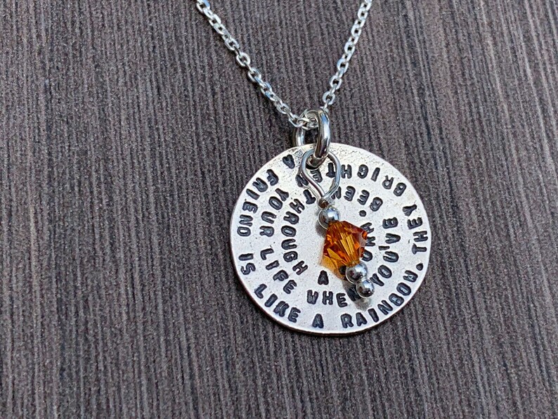 May include: Silver necklace with a round pendant that reads "The rainbow you live through is like a friend and your life is enough". The pendant has a small amber crystal charm.