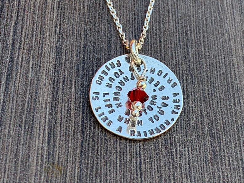 May include: Silver necklace with a round pendant that reads "They say you've been through a storm. When you live through your rainbow is like a friend to you". A red crystal hangs from the pendant.