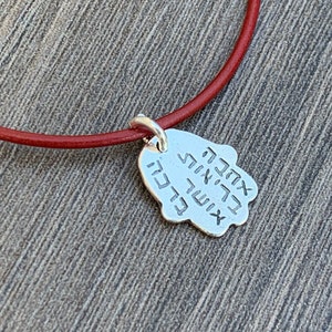 May include: A red leather cord necklace with a silver Hamsa hand charm. The charm has Hebrew writing on it.