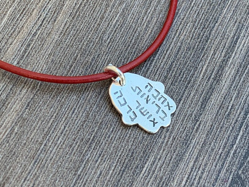 May include: A red leather cord necklace with a silver Hamsa hand charm. The charm has Hebrew writing on it.