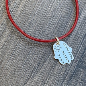 May include: A red leather bracelet with a silver charm in the shape of a hand. The charm has the words "LOVE", "HEALTH", "HAPPINESS" and a sun engraved on it.