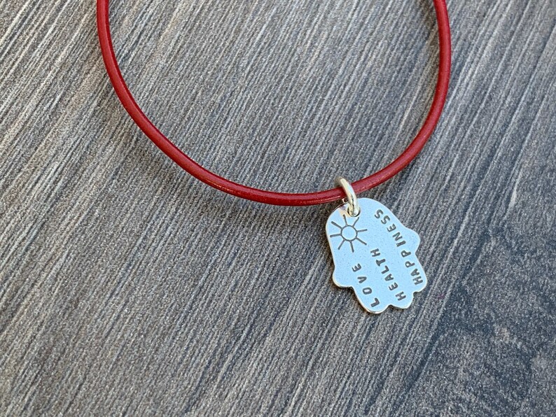 May include: A red leather bracelet with a silver charm in the shape of a hand. The charm has the words "LOVE", "HEALTH", "HAPPINESS" and a sun engraved on it.