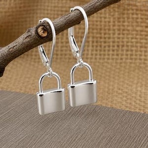 Op de afbeelding: Een paar zilverkleurige hangslot oorbellen. De oorbellen hangen aan een kleine, bruine tak. De achtergrond is een getextureerd, beige oppervlak en de oorbellen liggen op een grijs oppervlak.