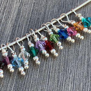 May include: A set of 12 colorful crystal stitch markers with silver beads and loops. The markers are attached to a silver wire.