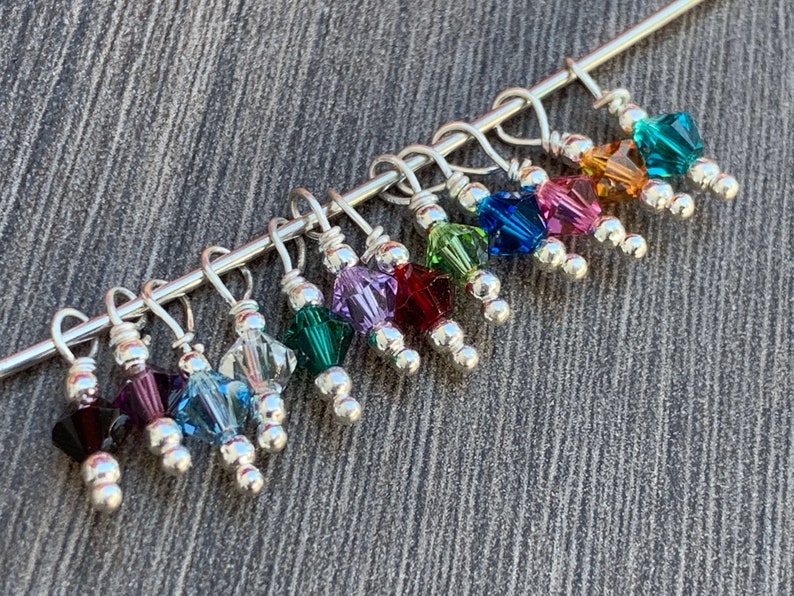 May include: A set of 12 colorful crystal stitch markers with silver beads and loops. The markers are attached to a silver wire.
