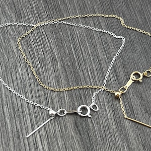 May include: Two delicate chains, one silver and one gold, are layered together. The chains have a simple, linked design and are connected by a small, round bead. The chains are displayed on a wooden surface.
