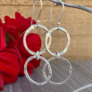 May include: A pair of silver-toned drop earrings featuring two interconnected circular hoops. Each hoop has a textured, hammered finish that reflects light. The earrings are suspended from simple silver-toned ear wires, creating a dangling design.