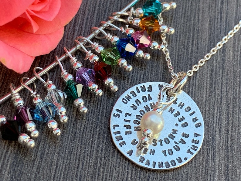 May include: A silver necklace with a charm that reads "A friend is like a rainbow. They brighten your life through every storm. When you've been through a storm, you give a rainbow." The charm is attached to a silver chain with a pearl bead. The necklace also has a silver bar with colorful crystal beads hanging from it.