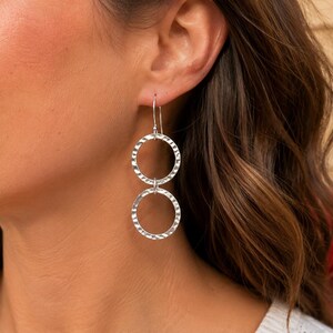 May include: A pair of silver drop earrings featuring two interconnected circular rings. Each ring has a textured, hammered finish that reflects light. The earrings are suspended from a simple silver hook wire that passes through the earlobe.