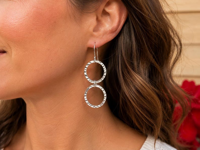 May include: A pair of silver drop earrings featuring two interconnected circular rings. Each ring has a textured, hammered finish that reflects light. The earrings are suspended from a simple silver hook wire that passes through the earlobe.