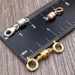 Op de afbeelding: Twee magnetische sluitingen voor kettingen of armbanden. Eén is zilverkleurig met een karabijnhaak en de andere is goudkleurig met een ronde ringsluiting. Beide sluitingen worden getoond naast een liniaal met inchmarkeringen.