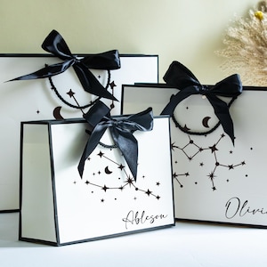 Könnte beinhalten: Weiße Geschenktüten mit schwarzen Rändern und Sternbildmustern. Jede Tasche ist mit einer schwarzen Satinschleife verziert. Die Namen "Ableson" und "Olivia" sind auf zwei der Taschen geschrieben. Verschiedene Größen.