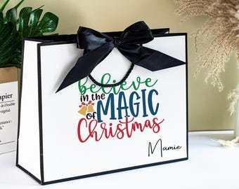 Puede incluir: Una bolsa de regalo blanca con un lazo y asas negras. La bolsa presenta el texto "believe in the MAGIC of Christmas" en verde, azul y rojo, con campanas doradas. El nombre "Mamie" está escrito en negro.