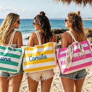 May include: Three women on a beach each carry a striped tote bag. The bags are green, yellow, and pink with white stripes. Each bag has a name embroidered in a contrasting color: "Luna", "Camila", and "Harley". The bags have rope handles.