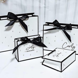 Pode incluir: Quatro sacolas de presente brancas com acabamentos e laços pretos. As sacolas são decoradas com pequenas pérolas falsas espalhadas. Duas sacolas têm nomes impressos e uma diz "Aly Bridesmaid".