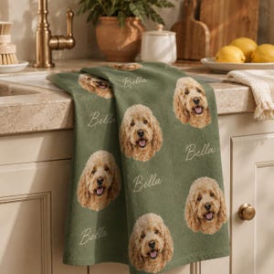 Pode incluir: Um pano de prato verde-sálvia com um padrão repetitivo do rosto de um cão de cor dourada e o nome Bella escrito em letra cursiva branca. O pano está sobre uma bancada de cozinha, exibindo a estampa detalhada do retrato do animal.