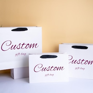 Może przedstawiać: Trzy białe torby na prezenty o różnych rozmiarach z czarnymi uchwytami. Torby mają napis "Custom" kursywą i słowa "gift bag" wydrukowane poniżej. Torby znajdują się na białej powierzchni.