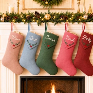 Peut inclure: Cinq chaussettes de Noël texturées en rose poudré, bleu clair, vert forêt, rose vif et rouge foncé. Chaque chaussette présente un revers brodé d'un prénom et d'une petite couronne avec un nœud rouge. Les prénoms sont Mom, Dad, Emma, Noah et Baby.