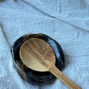 May include: A wooden spoon with a distinct grain pattern resting inside a small, round, black ceramic dish. The dish features an irregular, wavy rim and a glossy finish that contrasts with the natural, light brown texture of the wooden spoon.