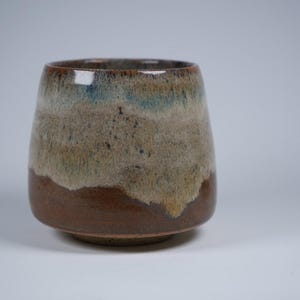 May include: A ceramic tumbler with a distinctive layered glaze. The base is a deep brown, fading to tan and green, with a hint of blue at the rim. The cup has a rounded form and a solid base. Ideal for beverages.