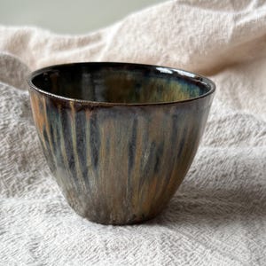 May include: A ceramic bowl with a tapered shape and a smooth rim. The exterior features a reactive glaze with vertical streaks of dark brown, deep blue, and earthy ochre tones that blend together to create a multi-coloured, layered appearance.