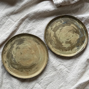 May include: Two circular metal plates featuring a distressed, brushed brass finish with visible swirling brush strokes in shades of muted gold, olive, and dark brown. Each plate has a thin, raised rim and a flat, circular base surface.