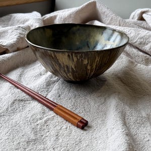 May include: A ceramic bowl with a mottled, dark green and brown glaze featuring vertical drip patterns, paired with a set of dark reddish-brown wooden chopsticks that have a textured, light brown wrapped detail near the handle end.