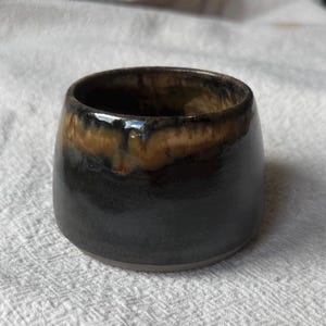 Può includere: Una piccola tazza in ceramica arrotondata con finitura lucida, caratterizzata da una base nero profondo che sfuma in uno smalto color marrone miele caldo vicino al bordo. Il recipiente presenta una silhouette liscia e curva.