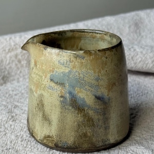Op de afbeelding: Een kleine keramische melkkan met een afgerond lichaam en een korte schenktuit. De buitenkant is voorzien van een gevlekt glazuur in aardse tinten beige, bruin en gedempt blauw, met een getextureerde, onregelmatige afwerking over het hele oppervlak.