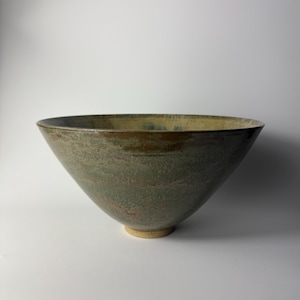 May include: A large, handmade ceramic bowl with a wide, open top and a conical shape. The bowl has a muted green and brown colour palette with a textured surface. The base is a lighter tan colour. This decorative bowl is suitable for serving or display.
