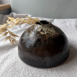 May include: A low, rounded ceramic vase with a wide base and a narrow opening. The exterior features a dark brown, glossy glaze that transitions into a mottled cream and white pattern near the top rim. A single dried, light beige plant stem is inserted.