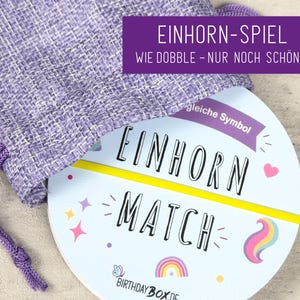 Könnte beinhalten: Ein violetter Stoffbeutel mit Kordelzug enthält ein rundes Kartenspiel mit der Aufschrift Einhorn Match. Die Karte zeigt den Text Einhorn Match, eine gelbe horizontale Linie, einen Regenbogen, ein rosa Herz, Sterne und Birthdaybox.de.