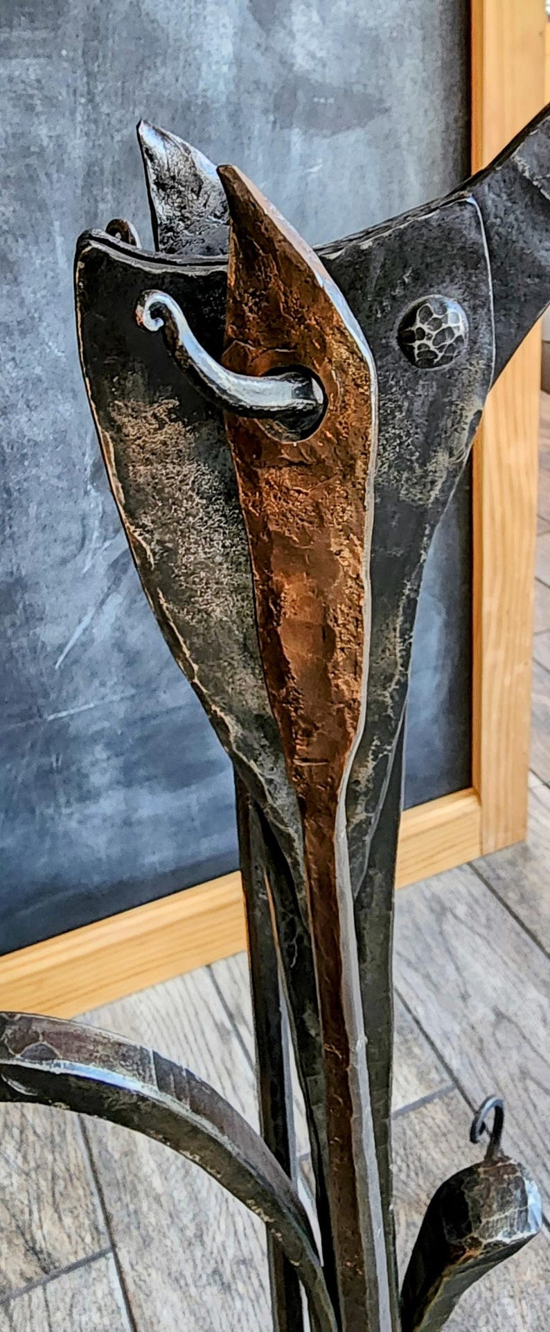 May include: A metal sculpture of a stylized plant with a copper-colored stem and black metal leaves. The sculpture has a rustic, hand-forged look.